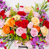A bountiful and cheerful floral arrangement in a clear glass vase. The bouquet is a vibrant mix of colorful roses in shades of red, peach, cream, and pink, interspersed with small red carnations and purple filler flowers. The arrangement is accented with various green sprigs and foliage, adding texture and depth. A light pink satin ribbon is tied around the vase. This bright and joyful bouquet is a perfect centerpiece for any celebration, expressing happiness and affection.
