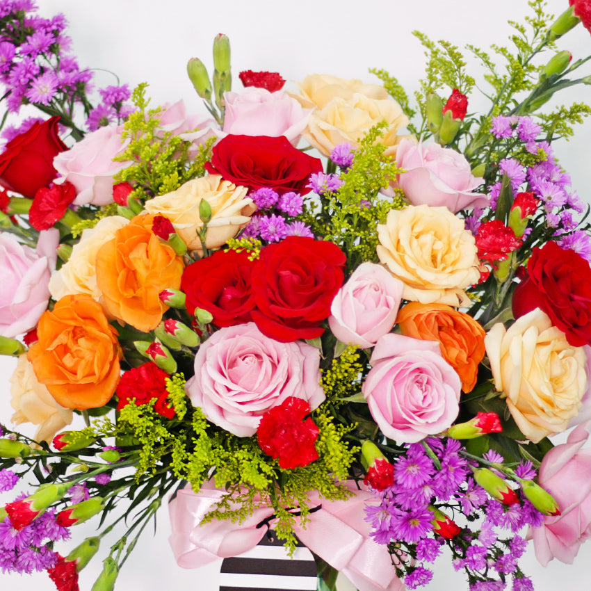 A bountiful and cheerful floral arrangement in a clear glass vase. The bouquet is a vibrant mix of colorful roses in shades of red, peach, cream, and pink, interspersed with small red carnations and purple filler flowers. The arrangement is accented with various green sprigs and foliage, adding texture and depth. A light pink satin ribbon is tied around the vase. This bright and joyful bouquet is a perfect centerpiece for any celebration, expressing happiness and affection.