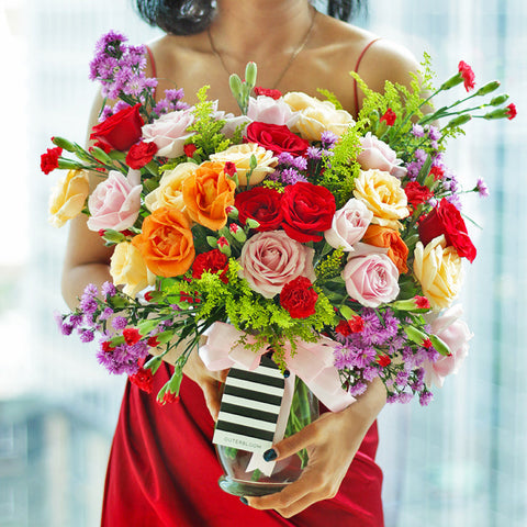 A bountiful and cheerful floral arrangement in a clear glass vase. The bouquet is a vibrant mix of colorful roses in shades of red, peach, cream, and pink, interspersed with small red carnations and purple filler flowers. The arrangement is accented with various green sprigs and foliage, adding texture and depth. A light pink satin ribbon is tied around the vase. This bright and joyful bouquet is a perfect centerpiece for any celebration, expressing happiness and affection.