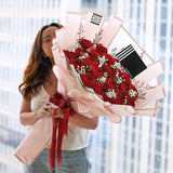 A luxurious and romantic bouquet featuring a generous cluster of approximately 20 red roses. The deep red roses are accented with delicate white baby's breath and thin sprigs of dried filler flowers. The bouquet is wrapped in a soft, semi-transparent pink paper with a subtle striped texture. A double red ribbon is tied into a beautiful bow at the base, and the bouquet includes a stylish black tag. This elegant arrangement is the perfect expression of love and passion.