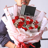 A luxurious and romantic bouquet featuring a generous cluster of red roses. The deep red roses are accented with delicate white baby's breath and thin sprigs of dried filler flowers. The bouquet is wrapped in a soft, semi-transparent pink paper with a subtle striped texture. A double red ribbon is tied into a beautiful bow at the base, and the bouquet includes a stylish black tag. This elegant arrangement is the perfect expression of love and passion.