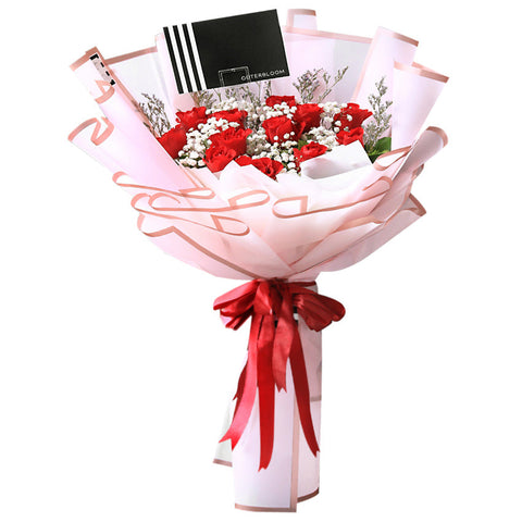A luxurious and romantic bouquet featuring a generous cluster of approximately 20 red roses. The deep red roses are accented with delicate white baby's breath and thin sprigs of dried filler flowers. The bouquet is wrapped in a soft, semi-transparent pink paper with a subtle striped texture. A double red ribbon is tied into a beautiful bow at the base, and the bouquet includes a stylish black tag. This elegant arrangement is the perfect expression of love and passion.