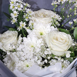 An elegant bouquet featuring a monochromatic white and grey theme. The arrangement is composed of classic white roses, asters, and peacock, delicate baby's breath, and other smaller white filler flowers. The flowers are wrapped in a clean, white inner layer and a sophisticated grey outer layer. The bouquet is tied with a double white ribbon—and a small tag. The minimalist style makes it a thoughtful gift for any occasion.